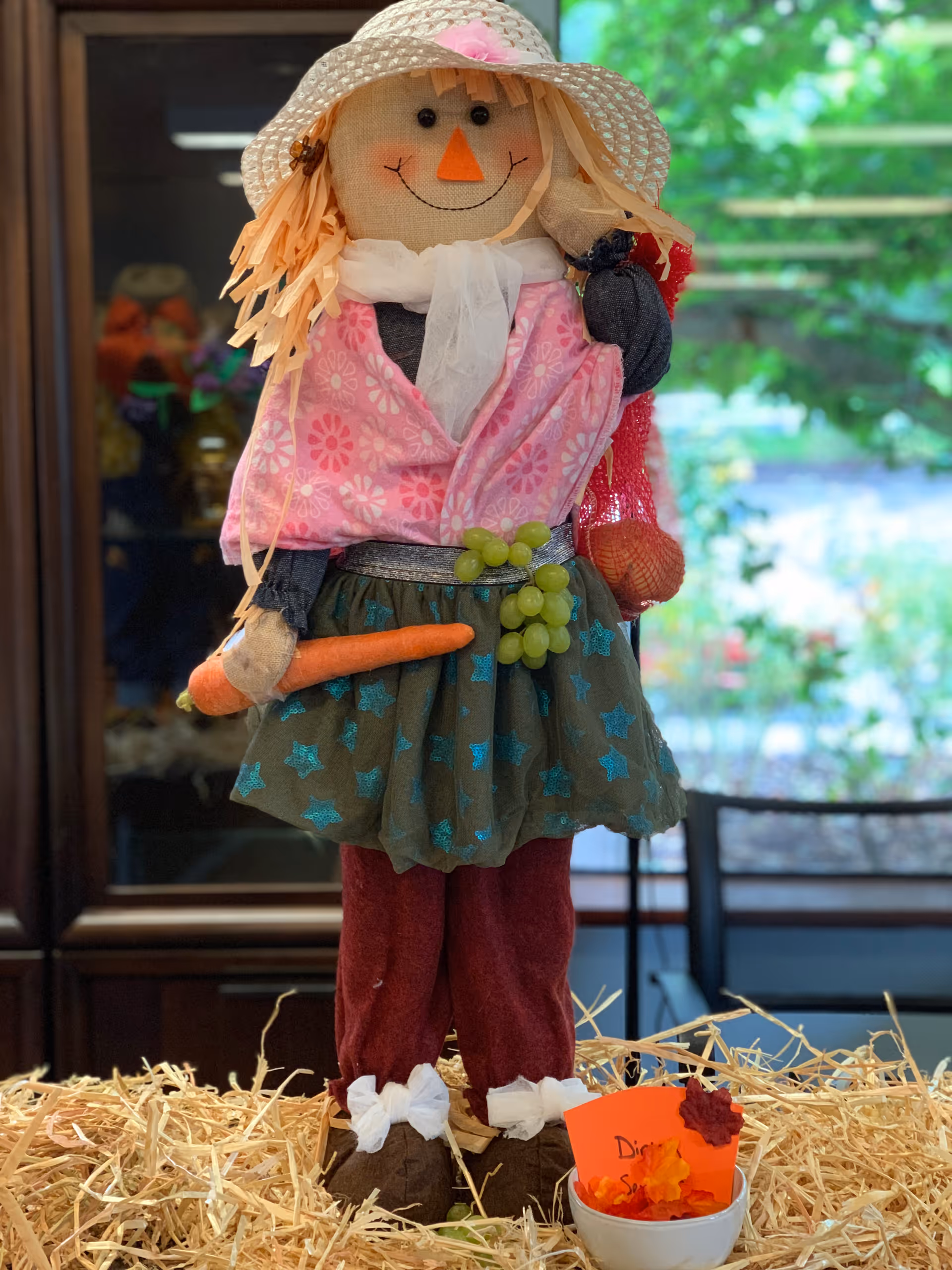 A decorative scarecrow figure wearing a straw hat and pink jacket holds a carrot and grapes while standing on hay indoors.