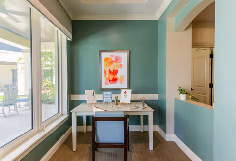 A small interior workspace with a white desk and a blue chair positioned in front of a green wall. On the desk are several decorative items, including framed pictures and a small plant. A large window to the left lets in natural light and shows an outdoor patio area with chairs and a table. The room has white trim and a partial wall with an open archway on the right side.
