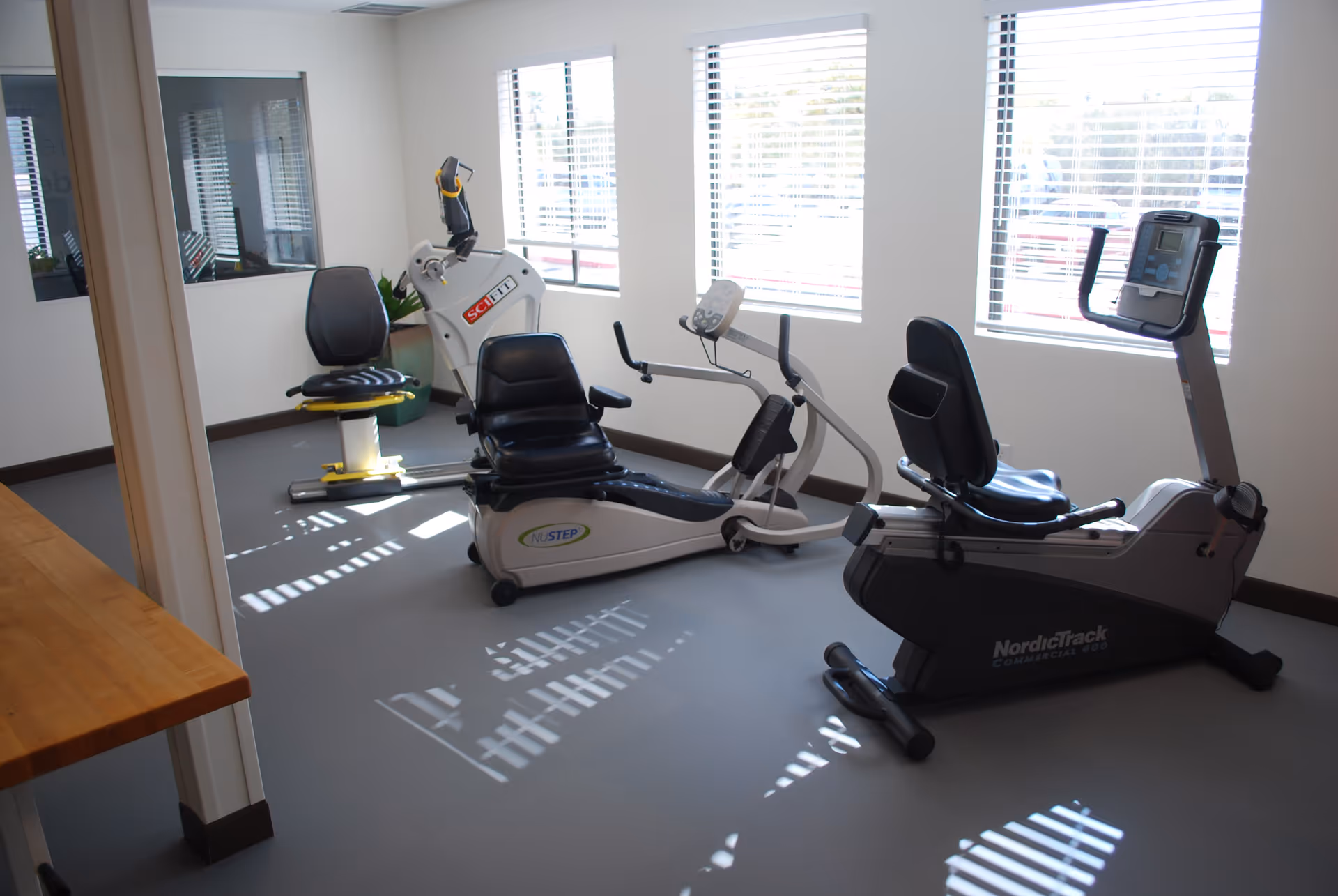 A small exercise room with three recumbent exercise bikes positioned near windows with blinds. The room has light-colored walls and a gray floor, with natural light coming through the windows.