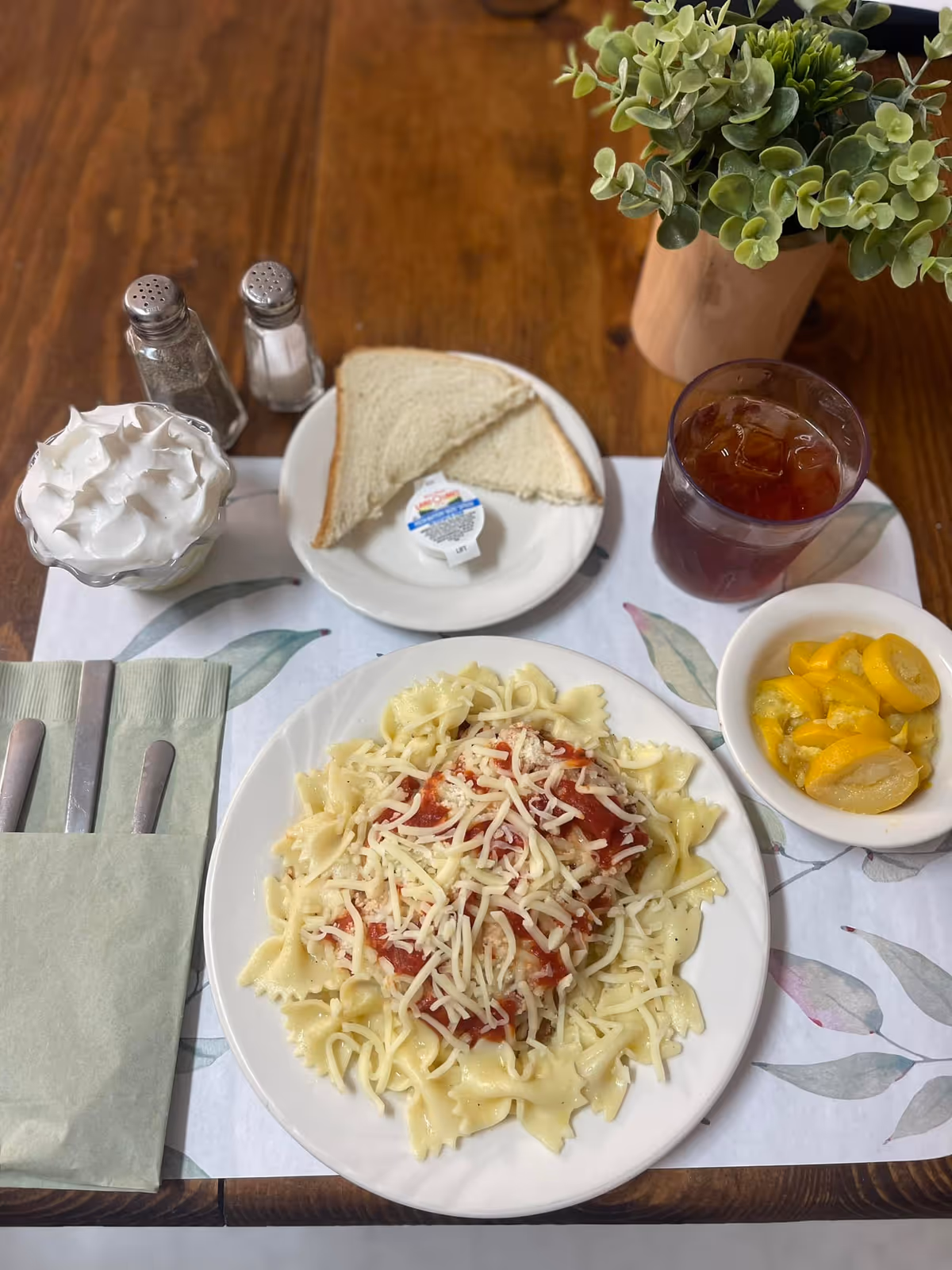 A meal served on a wooden table with a plate of bowtie pasta topped with marinara sauce and shredded cheese, a small bowl of sliced peaches, a glass of iced tea, a plate with two slices of white bread and a small container of margarine, a cup of whipped cream, salt and pepper shakers, and a set of utensils wrapped in a green napkin.