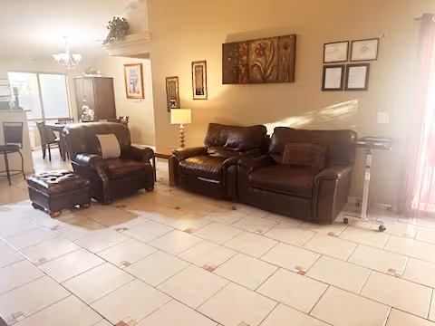 A well-lit living room with two dark brown leather sofas and a matching ottoman on a tiled floor. The room features a side table with a lamp, wall art, and framed certificates on the wall. In the background, there is a dining area with a table and chairs near a window.