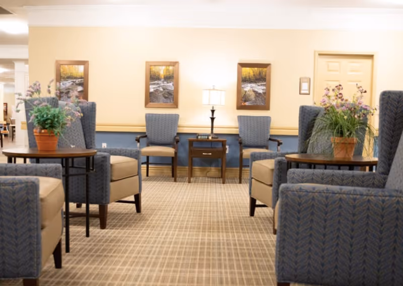 A cozy seating area in a senior living facility with multiple blue upholstered armchairs and wooden side tables. Two potted plants with flowers are placed on the tables. The walls are painted beige with a blue wainscot, and three framed pictures of a river scene hang on the wall. A table lamp is on a small wooden table against the wall, and a closed door is visible in the background.