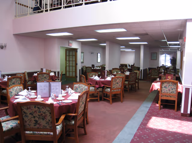 Interior view of a dining room with multiple round tables covered with maroon tablecloths, each set with white napkins, cups, and menus. The chairs have floral upholstery, and the room has a carpeted floor with a patterned runner. Fluorescent ceiling lights illuminate the space.