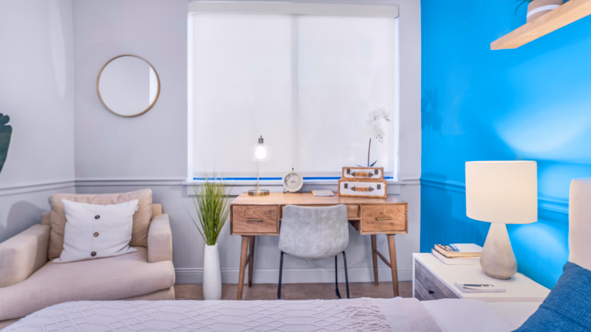 A cozy bedroom with a beige armchair with a white pillow on the left, a wooden desk with a gray chair in front of a window with white blinds, and a white nightstand with a lamp and books on the right. The wall behind the nightstand is painted bright blue, and there is a round mirror on the left wall.