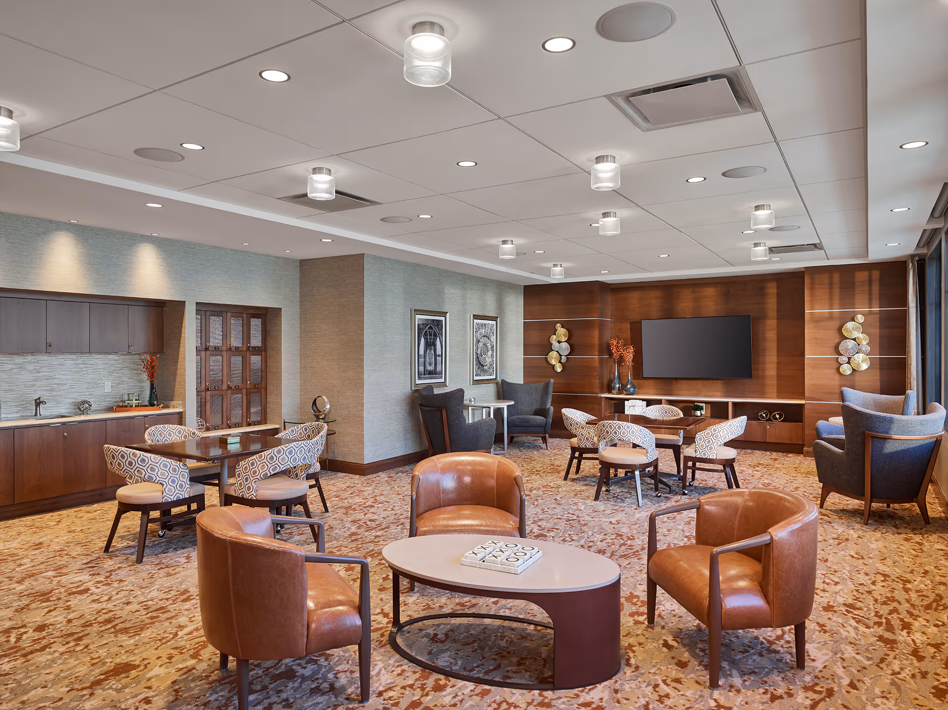 A spacious and well-lit common area with modern furniture including leather armchairs around a coffee table, patterned chairs around small tables, and a large flat-screen TV mounted on a wooden accent wall. The room features a carpeted floor, recessed lighting, and a small kitchenette area with cabinets and a sink.