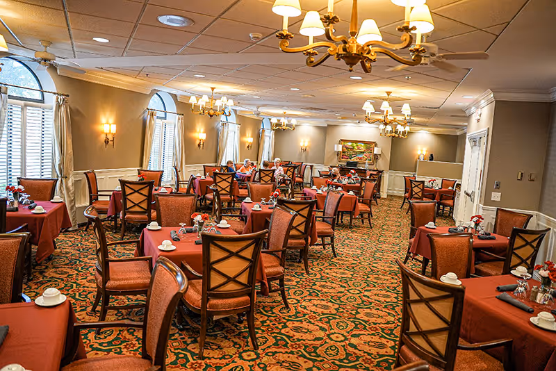 Spacious dining room with red-tablecloth tables and wooden chairs, chandeliers, patterned carpet, and a few seated residents.