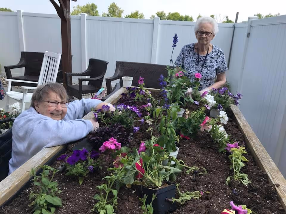 Two elderly women gardening together in a raised garden bed filled with soil and colorful flowers, surrounded by outdoor chairs and a white fence.