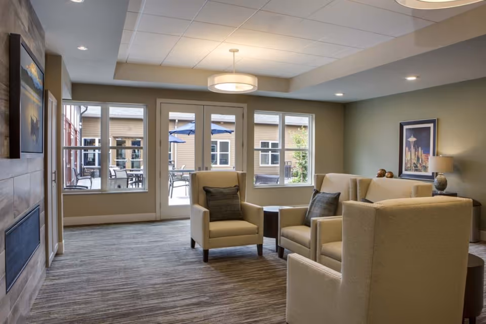 A cozy living room area with beige armchairs and a sofa arranged around a small round table. The room has large windows and glass doors that open to an outdoor patio with tables and umbrellas. The walls are painted green and beige, with framed artwork and a table lamp on a side table. The ceiling has recessed lighting and a modern circular light fixture.