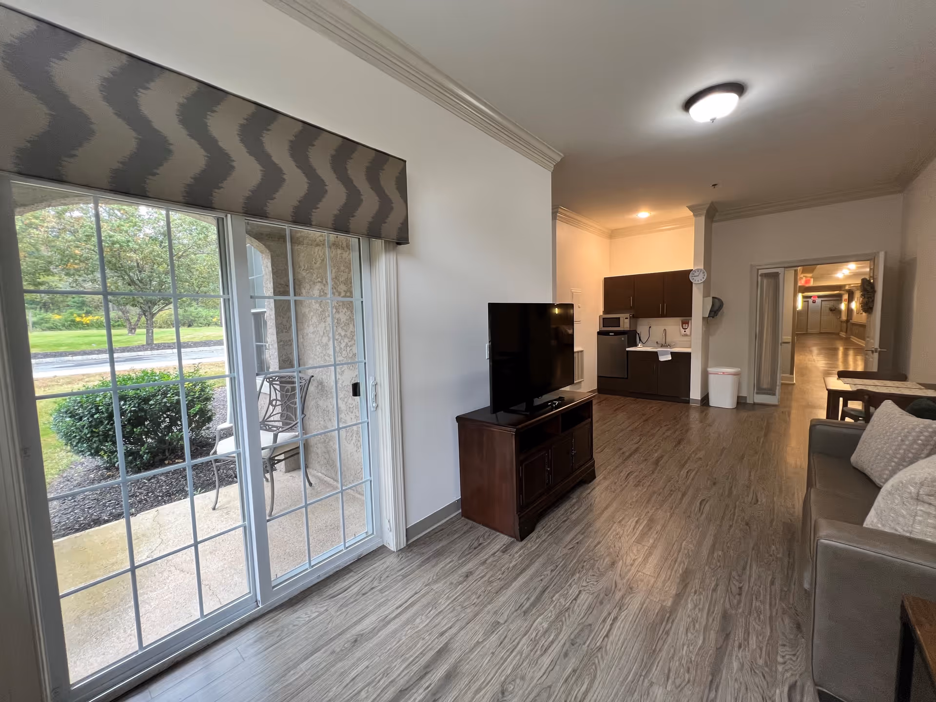 Interior view of a living area in an assisted living facility showing a sliding glass door leading to an outdoor patio with chairs. Inside, there is a TV on a wooden stand, a small kitchenette with cabinets, a microwave, and a sink. A couch with pillows is visible on the right, and a hallway extends into the background.