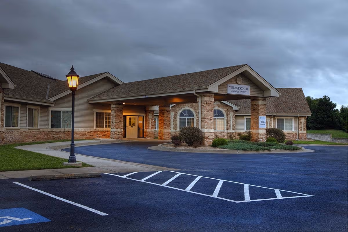 Front entrance of a single-story brick nursing facility with a covered drive-through, lit lamp post, landscaping, and an empty parking lot.