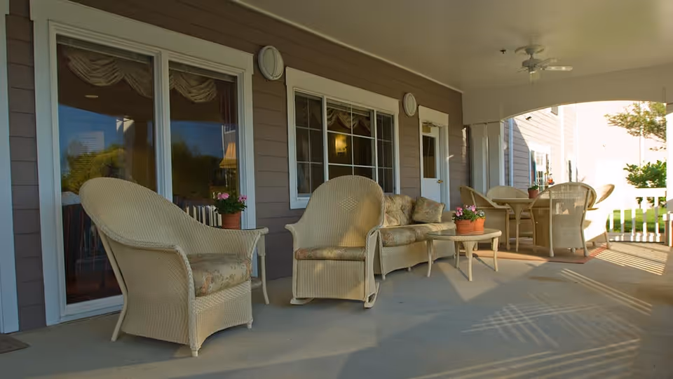 Covered outdoor patio area with wicker chairs, a loveseat, and tables with potted plants. The patio is attached to a building with large windows and a door, and there is a ceiling fan above.