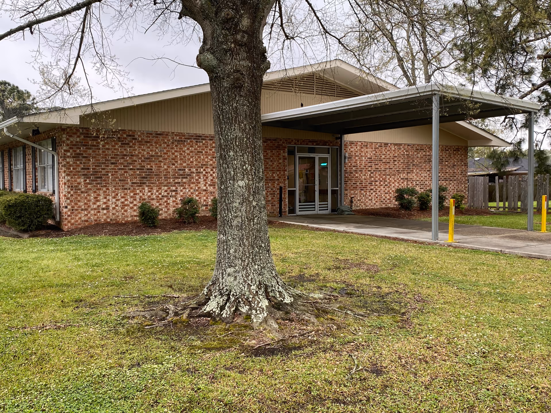Exterior view of a single-story brick building with a covered entrance supported by metal poles. There is a large tree in the foreground on a grassy lawn, and some small bushes are planted along the building's side. The sky is overcast.