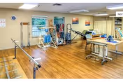 A physical therapy or rehabilitation room with wooden flooring, parallel bars for walking exercises, exercise equipment, therapy tables, and windows with blinds. The room is well-lit with ceiling lights and has some wall decorations.