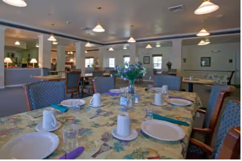 Dining room with a table set for a meal—plates, cups, utensils, and a vase of flowers—inside a memory care community.