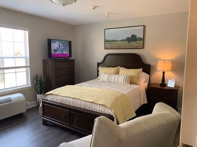 A cozy bedroom with a large bed featuring a dark wooden headboard and footboard, adorned with yellow and white bedding and pillows. A nightstand with a lamp and a small picture frame is on the right side of the bed. A tall dresser with a television on top is positioned against the wall near a window with white blinds. A beige armchair is partially visible in the foreground. The room has light-colored walls and dark wood flooring.