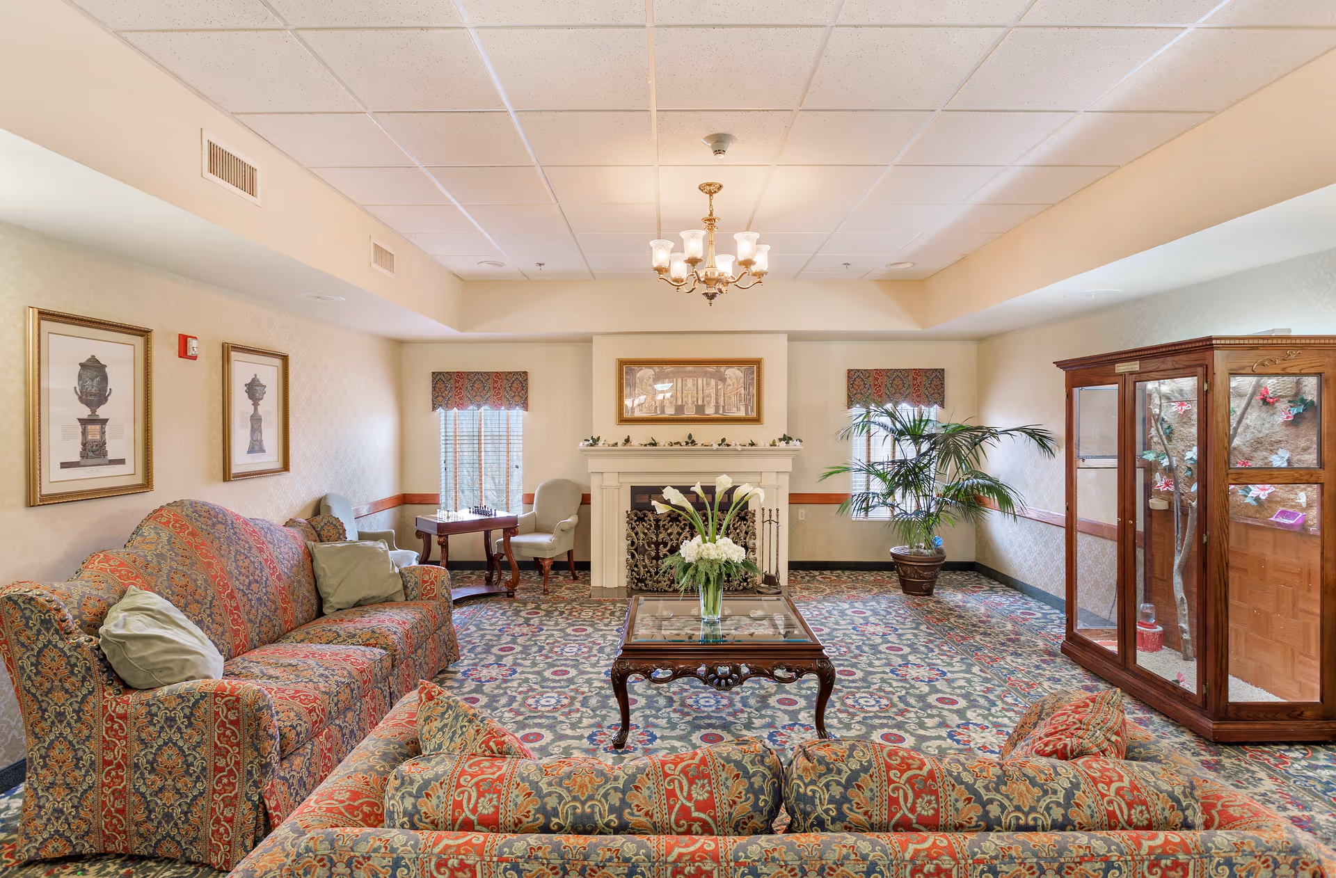 A cozy living room area in North Strabane Retirement Village featuring two patterned sofas with cushions, a wooden coffee table with a glass top and a vase of white flowers, a fireplace with decorative screen, two framed pictures on the wall, a small table with two chairs near the window, a large potted plant, and a wooden display cabinet with glass doors. The room has patterned carpet and soft lighting from a chandelier.