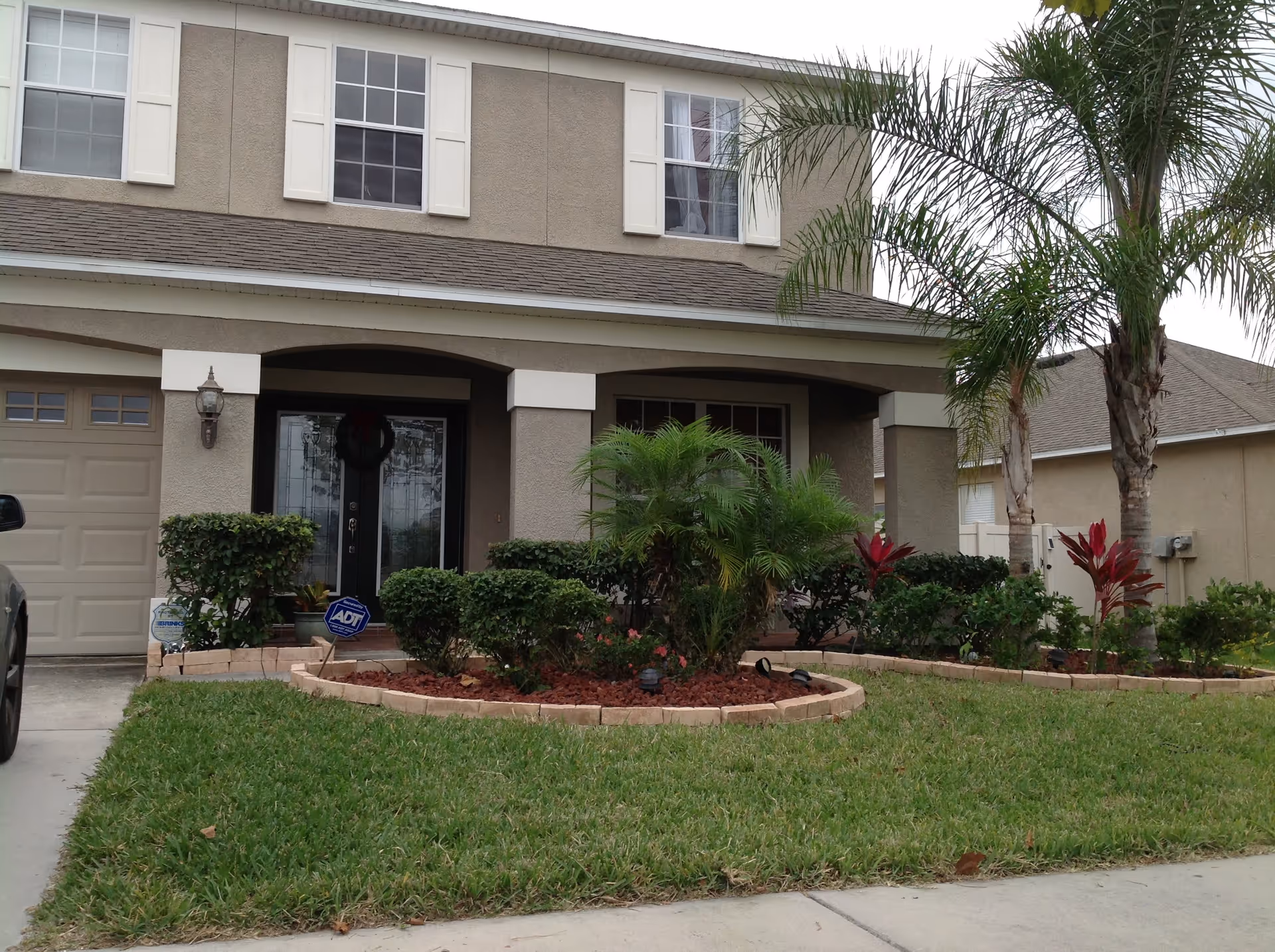 Front exterior view of a two-story residential building with beige walls, white window shutters, a dark front door with glass panels, a garage door, and a well-maintained garden with trimmed bushes, palm trees, and a green lawn.