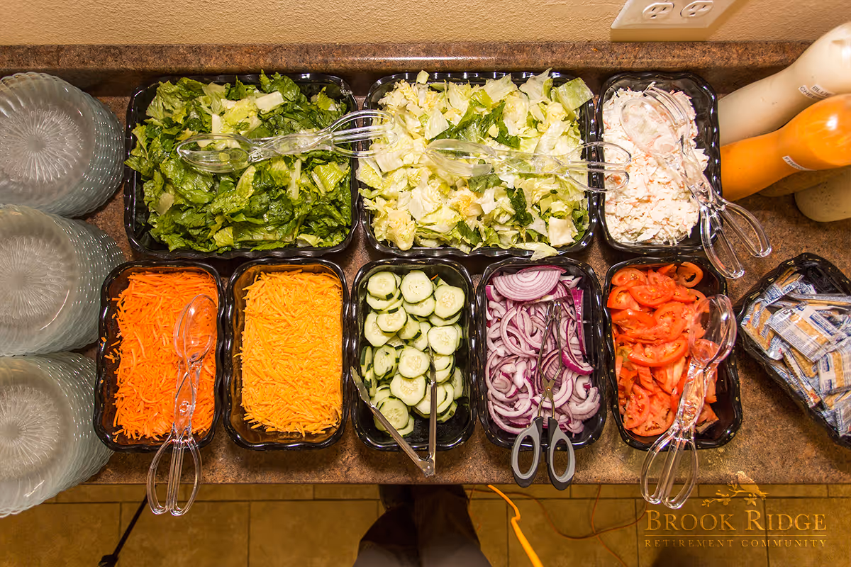A salad bar with various fresh vegetables and toppings including shredded carrots, shredded cheddar cheese, sliced cucumbers, sliced red onions, chopped tomatoes, two types of lettuce, and a creamy salad. There are clear plastic serving utensils in each container and stacks of clear plates on the left side. Bottles of salad dressings are visible on the right side.