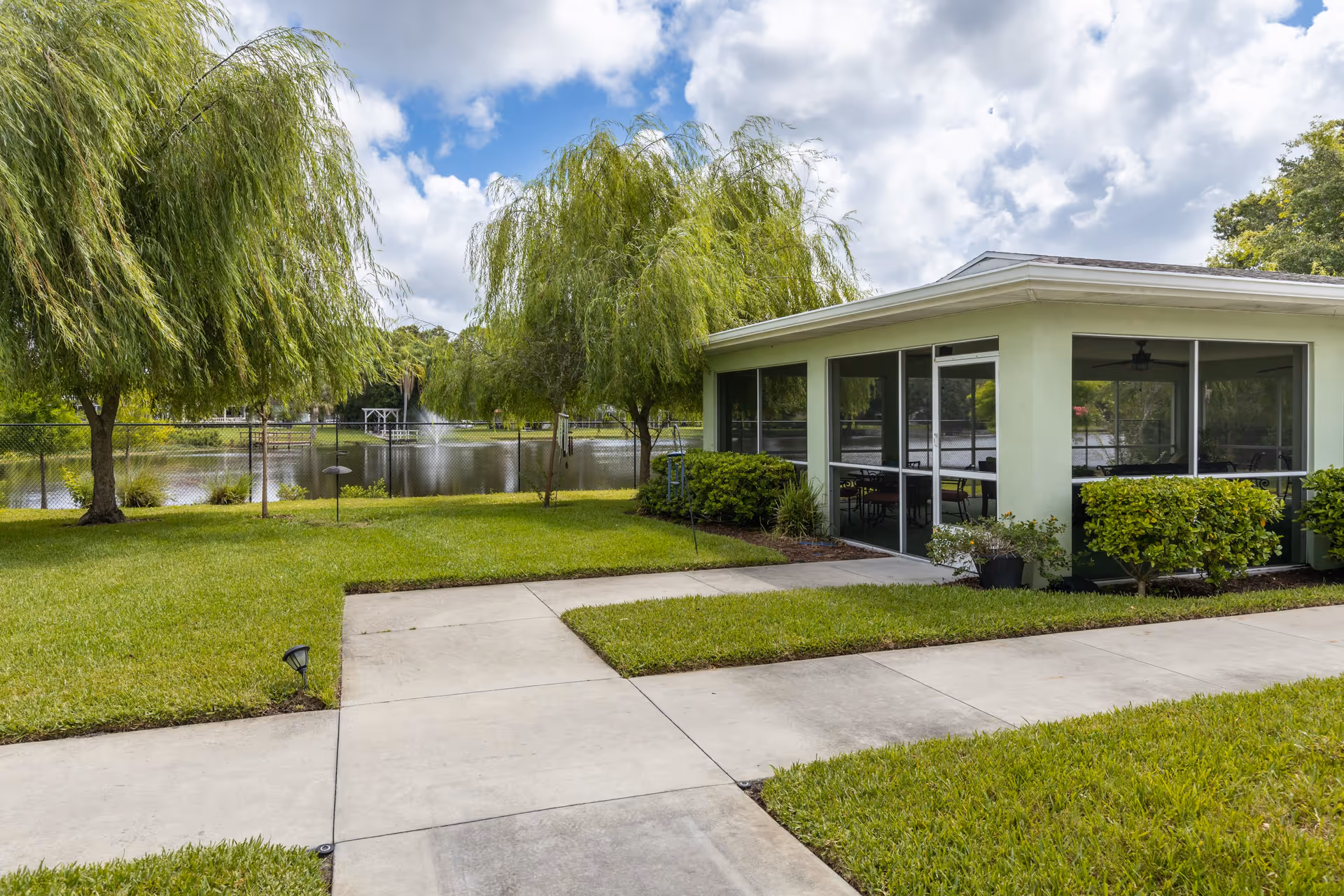 Screened patio of a single-story lakeside building with a concrete walkway, green lawn, weeping willow trees, and a pond with a fountain.