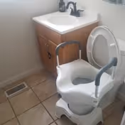A bathroom with a white toilet equipped with a raised commode chair with armrests. Next to the toilet is a wooden vanity with a white sink and a black faucet. The floor is tiled and there is a vent on the floor near the vanity.
