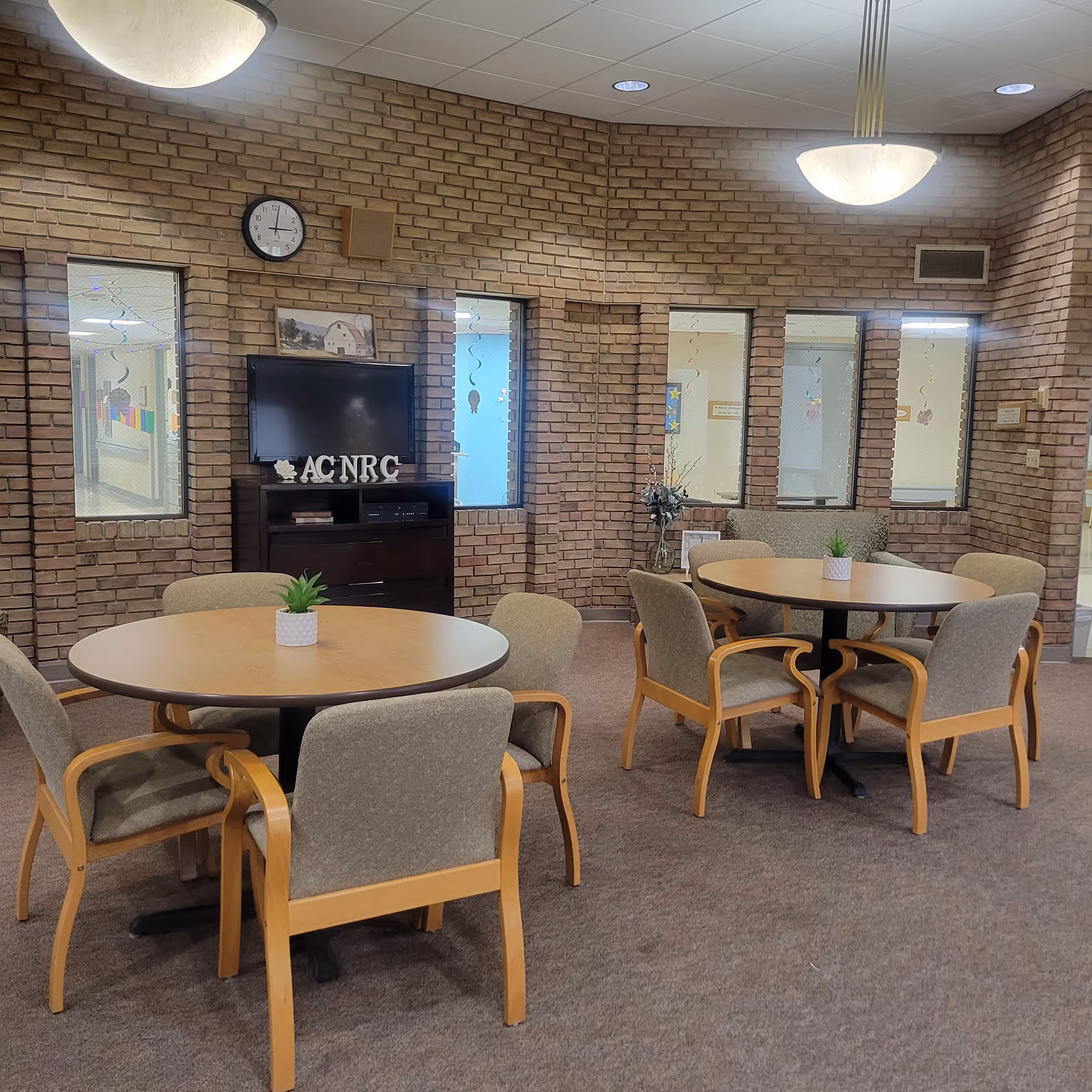A senior facility common room with round tables and chairs, a TV on a cabinet, and brick interior walls.