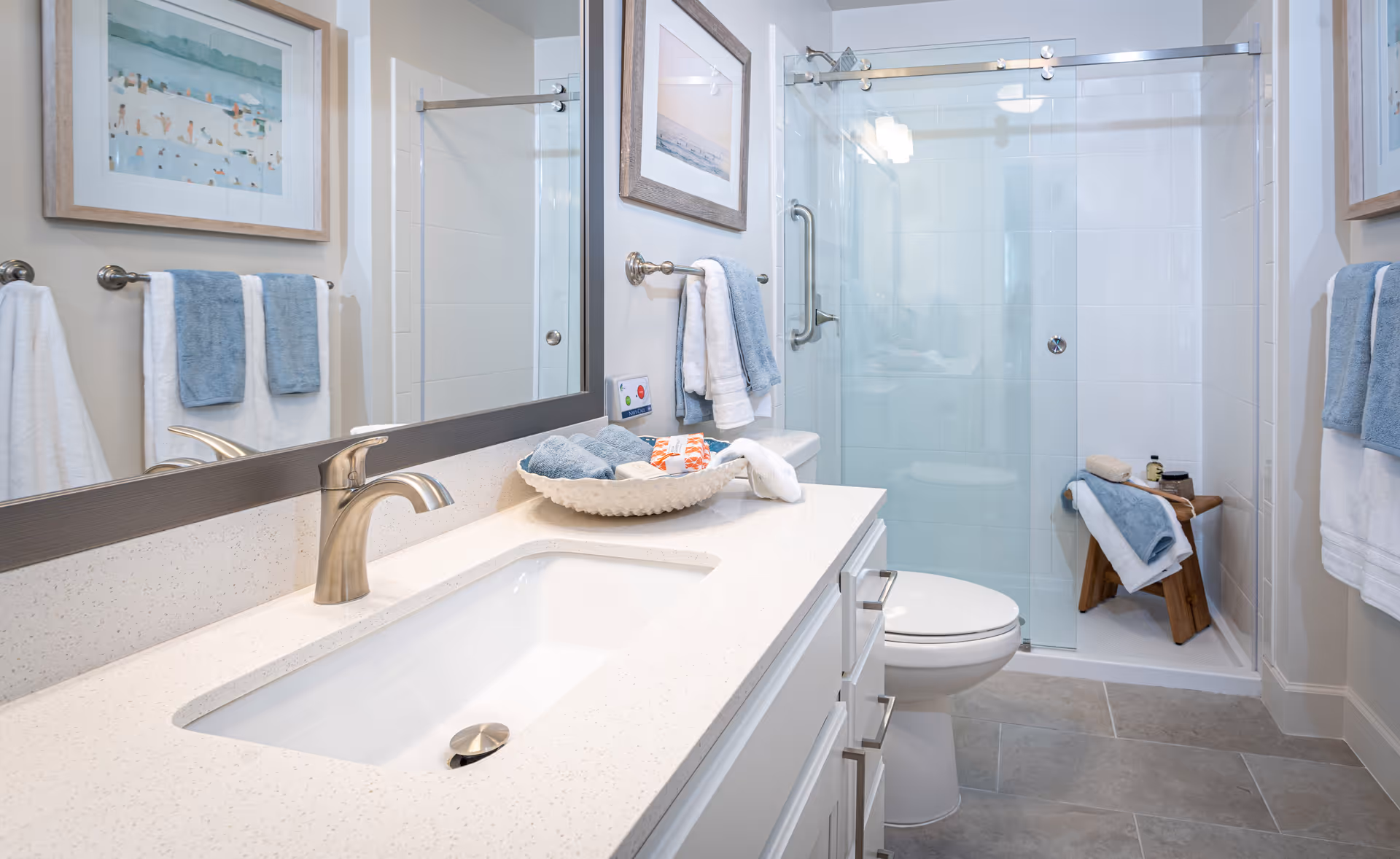 A clean and modern bathroom featuring a white countertop with an undermount sink and brushed nickel faucet. Above the sink is a large mirror reflecting the shower area with glass sliding doors. The shower has white tiled walls and a wooden stool with folded towels and toiletries. Blue and white towels hang on towel racks on the walls. The floor is tiled in a light gray color.