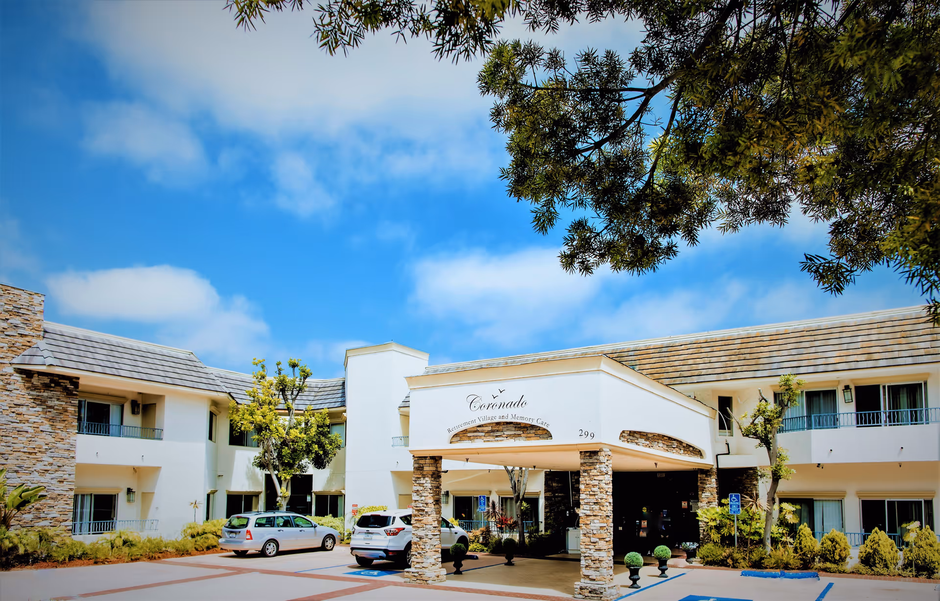 Front exterior view of Coronado Retirement Village building with a covered entrance supported by stone pillars, several parked cars, and trees under a blue sky with some clouds.