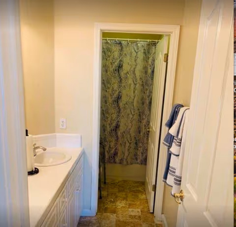 Small bathroom with a sink and vanity on the left, an open door revealing a shower with a patterned curtain, and towels hanging on the door.
