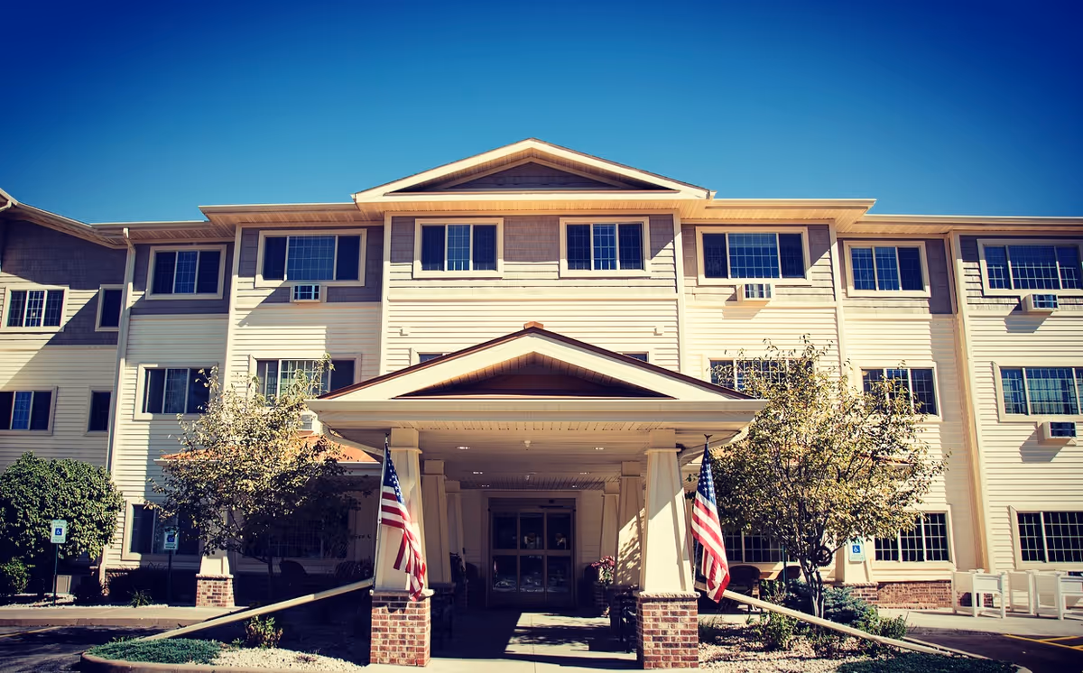 Front exterior view of a three-story assisted living facility building with beige siding and multiple windows. The entrance has a covered porch supported by columns with two American flags on either side. There are small trees and bushes near the entrance and a clear blue sky above.