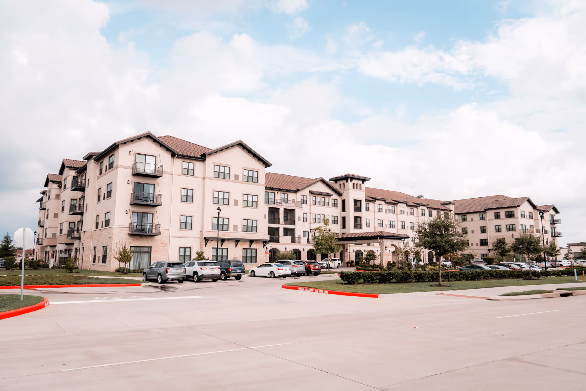 Large four-story senior living facility building with parked cars and landscaped entrance under a partly cloudy sky.