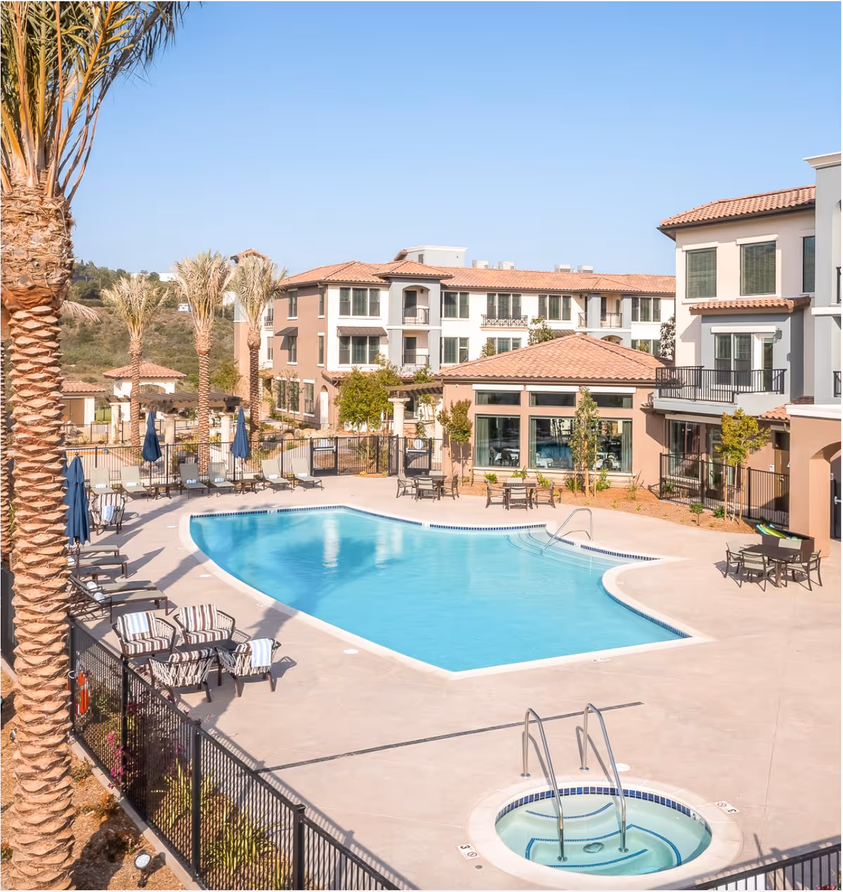 Outdoor swimming pool area at Santianna Oakmont Signature Senior Living with lounge chairs, umbrellas, palm trees, and a hot tub, surrounded by multi-story residential buildings under a clear blue sky.