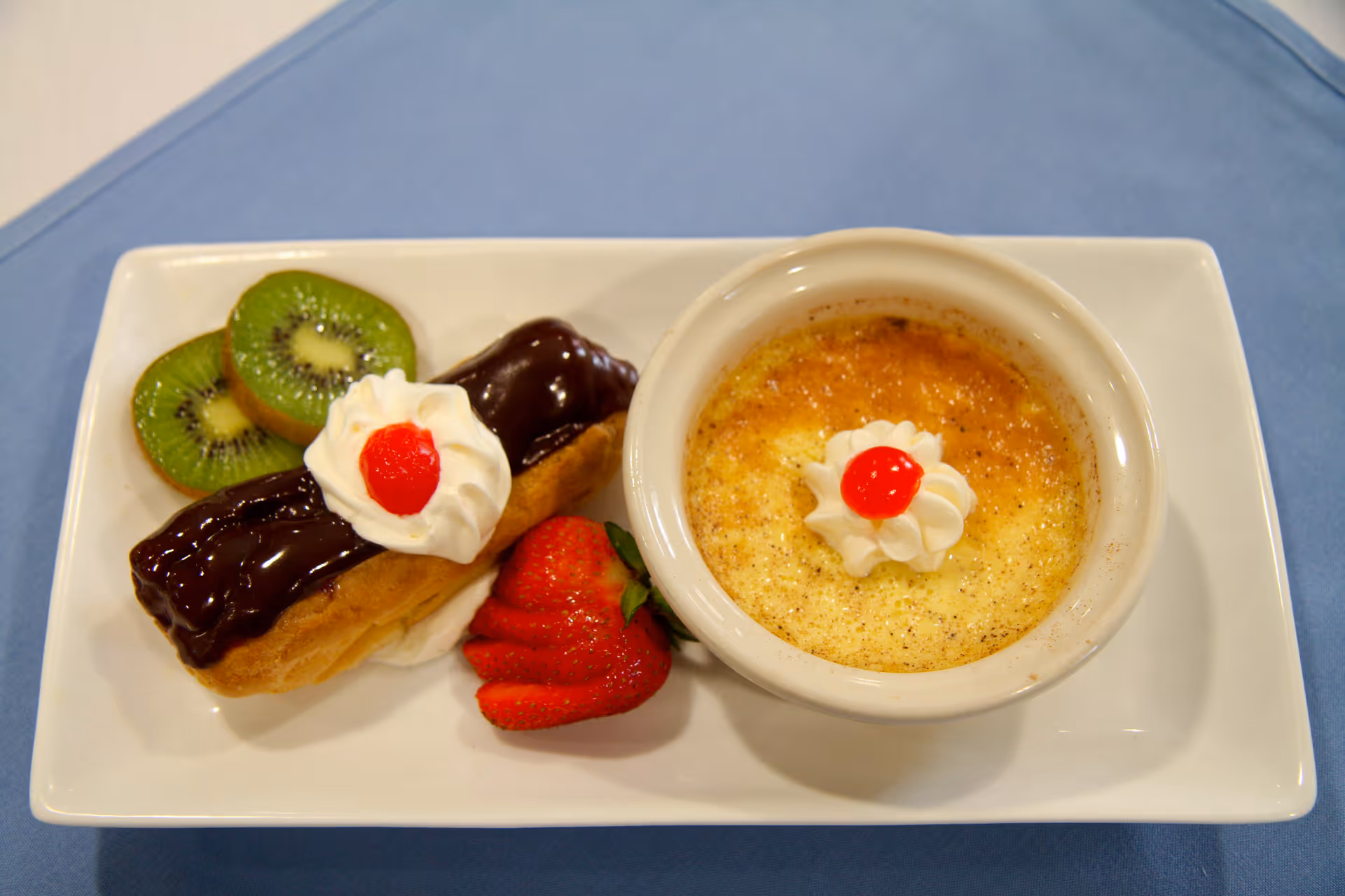 A dessert plate with a chocolate eclair topped with whipped cream and a cherry, fresh kiwi and strawberry slices, and a ramekin of crème brûlée garnished with whipped cream and a cherry.