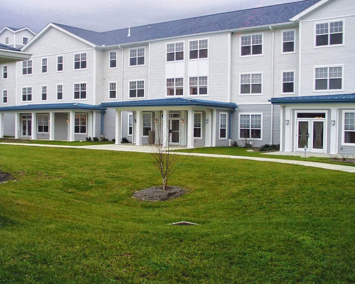 Front exterior of a three-story white senior living building with multiple windows, entrances, and a grassy lawn.