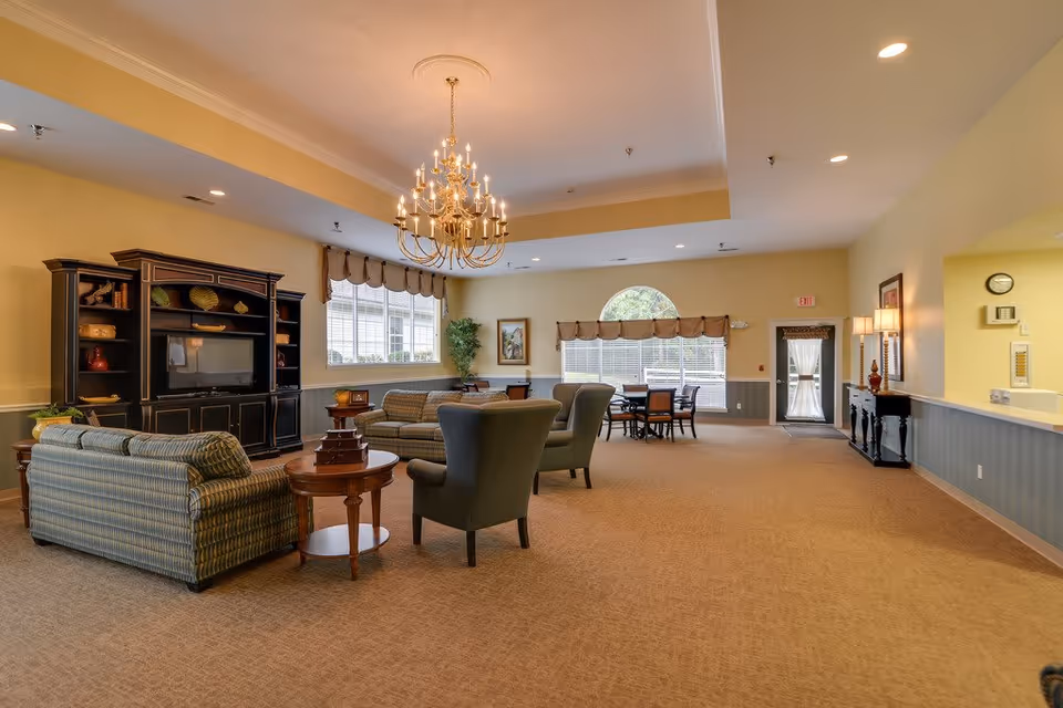 Spacious common lounge with upholstered chairs and sofas, a large entertainment cabinet, chandelier, and windows.