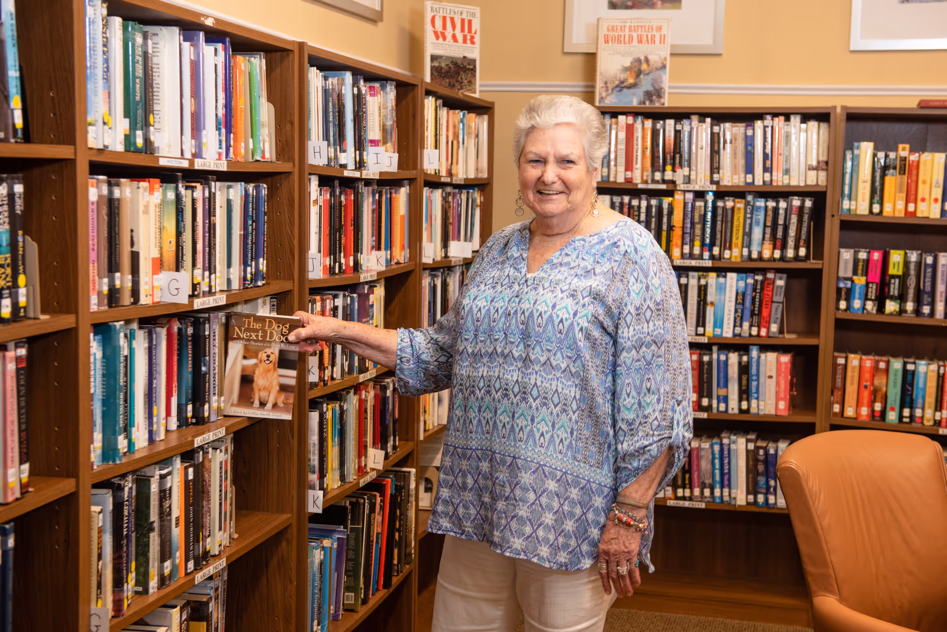 An elderly woman with short white hair wearing a blue patterned blouse and white pants is standing in a library. She is smiling and holding a book titled 'The Dog Next Door' while standing next to wooden bookshelves filled with books. The library has a warm, inviting atmosphere with framed pictures on the wall and a tan leather chair nearby.