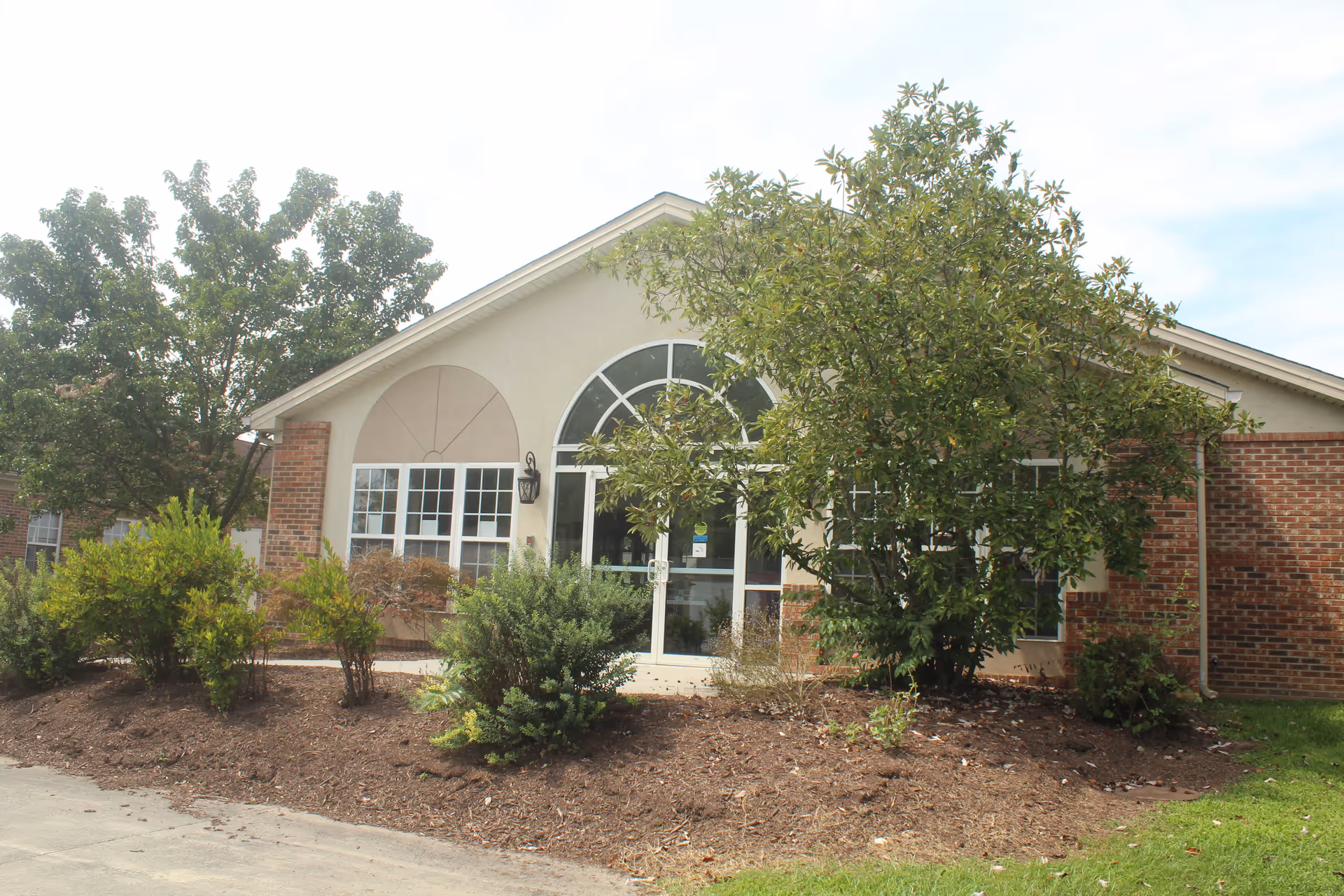 Exterior view of a single-story building with a peaked roof, large arched window above the entrance door, and several rectangular windows. The building is surrounded by landscaped bushes, small trees, and a mulched garden bed. The sky is partly cloudy.