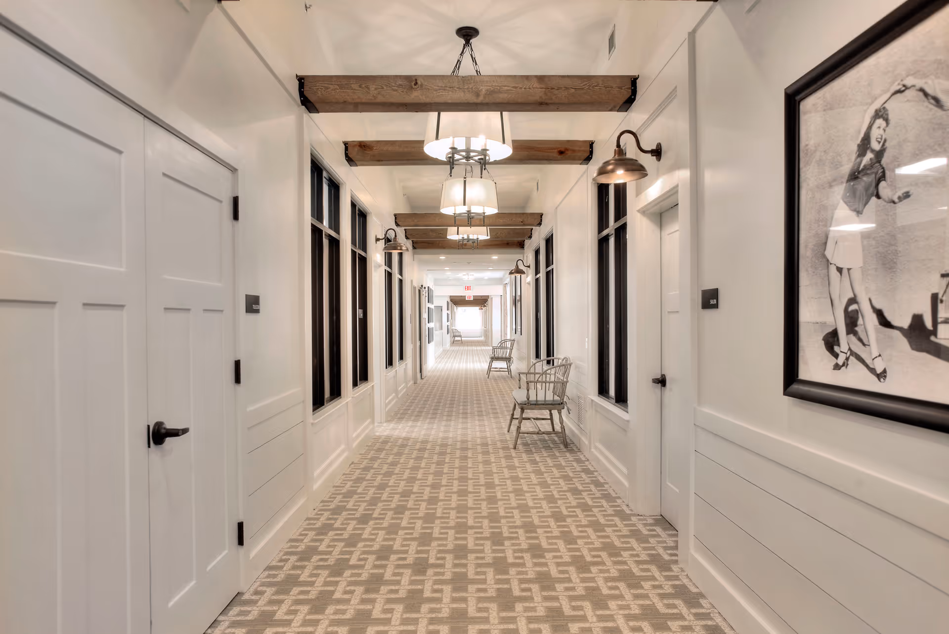 A bright, carpeted interior corridor with doors and windows along the sides, wooden ceiling beams, hanging light fixtures, and chairs placed against the walls.