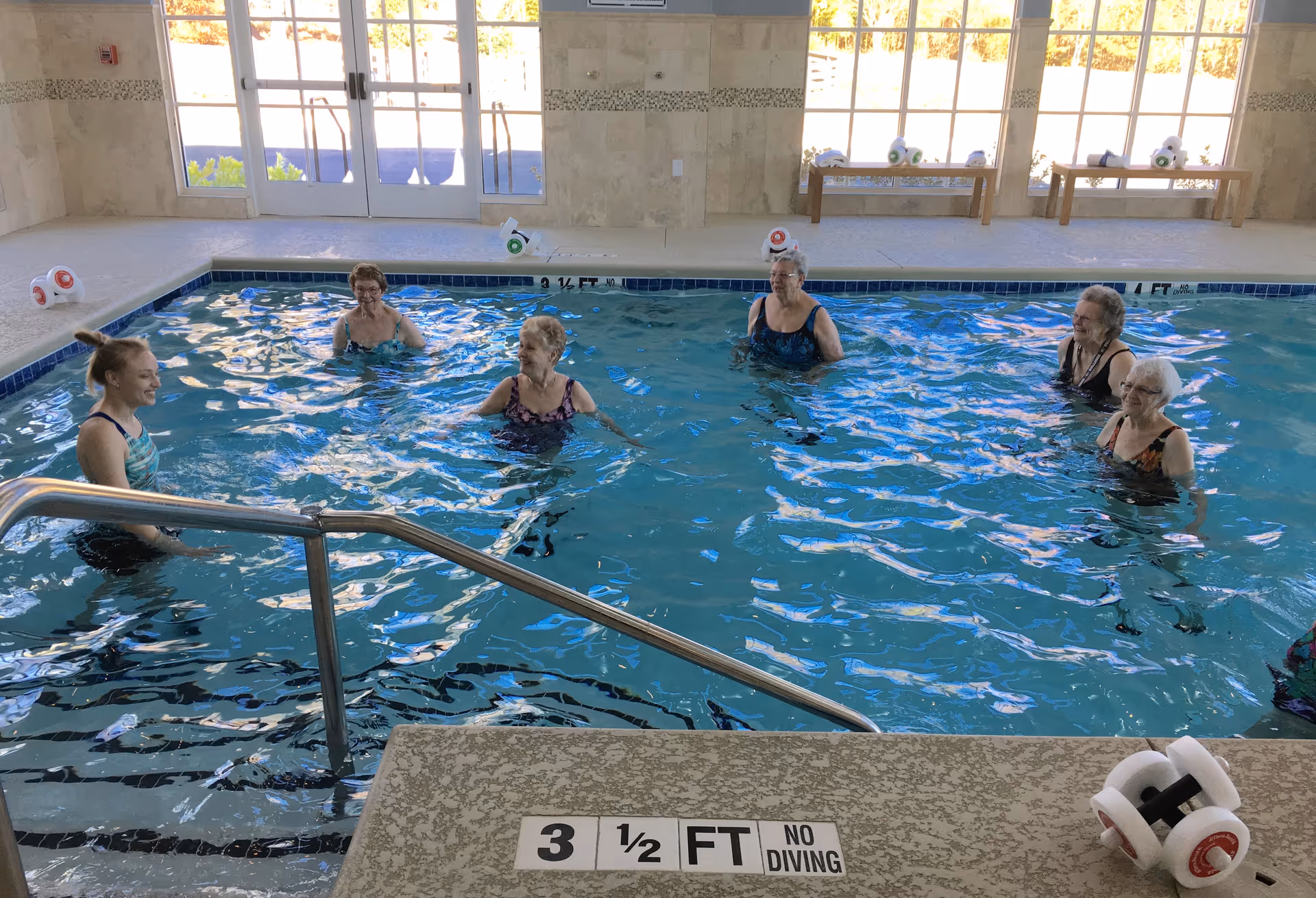 Six elderly women standing and smiling in an indoor swimming pool with clear blue water. The pool area has large windows letting in natural light, beige tiled walls, and a metal handrail leading into the water. There are small white and red pool weights placed around the pool edge.