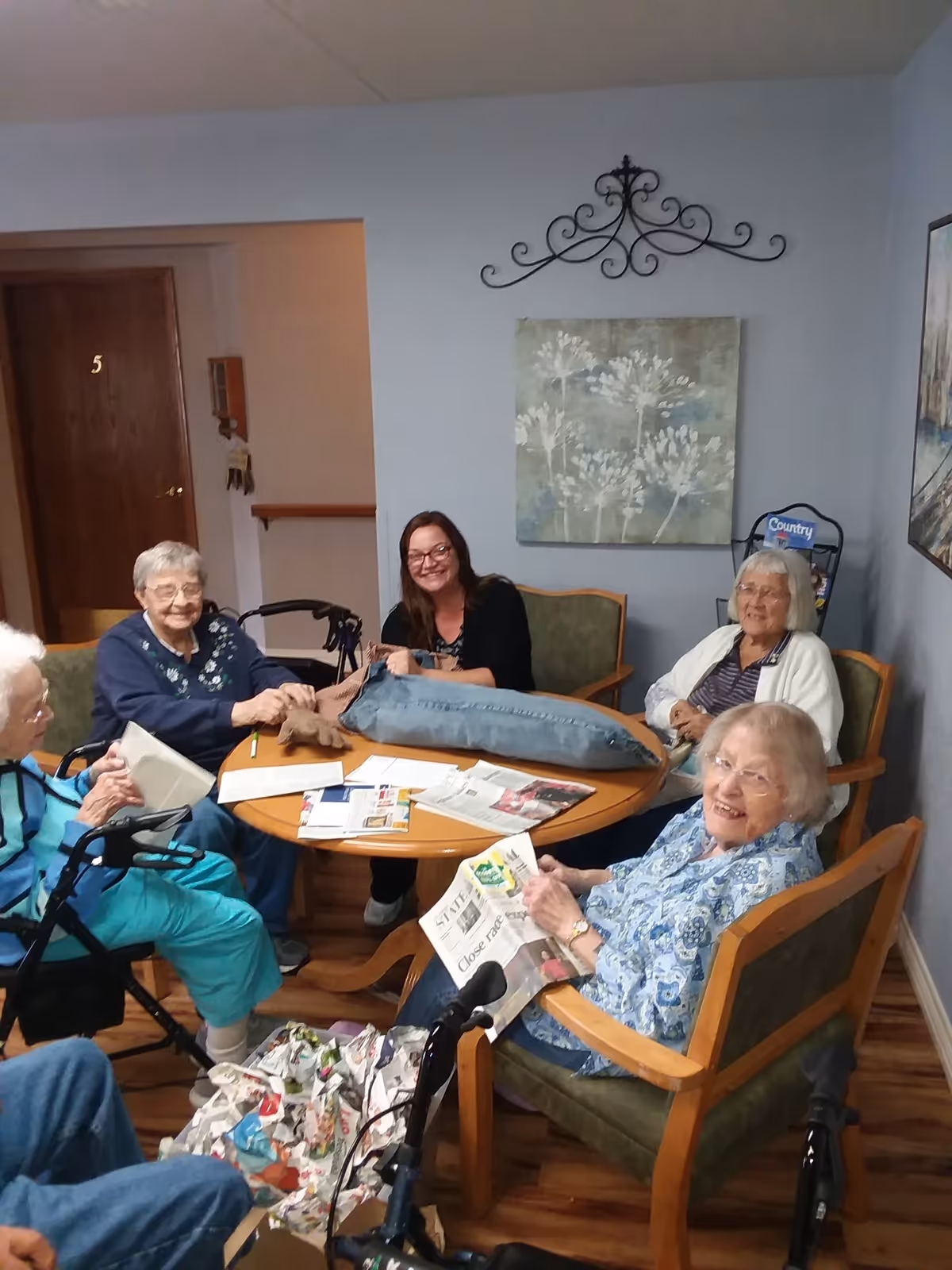 A group of elderly residents and a staff member sit around a round table in a cozy common room, reading newspapers and smiling.
