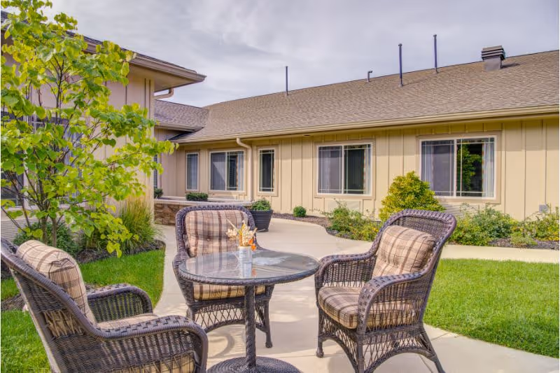 Outdoor patio area at Whitetail Springs Memory Care Community with a round glass-top table and four cushioned wicker chairs on a concrete path surrounded by green grass, plants, and a beige building with several windows in the background under a cloudy sky.