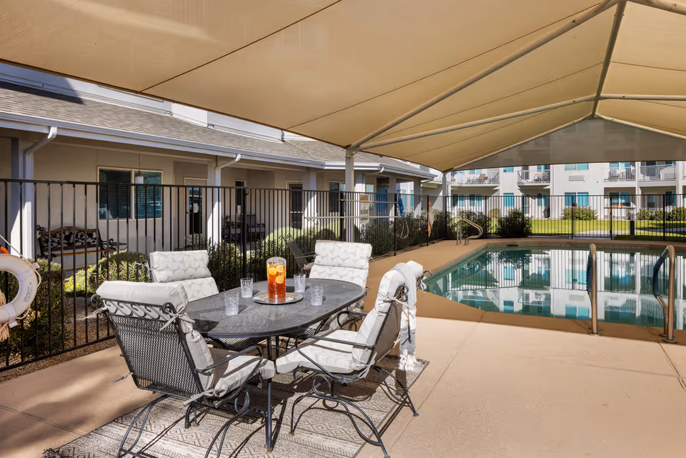 Shaded outdoor seating area with a table and four cushioned chairs next to a fenced swimming pool at a senior living community. A pitcher of iced tea and four glasses are on the table. The pool area is surrounded by buildings with balconies and windows.
