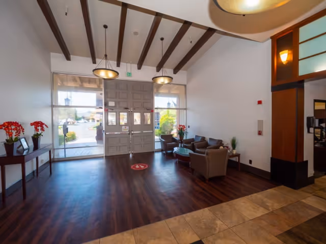 A spacious lobby area with wooden flooring and a tiled entrance. There are large double doors with glass panels letting in natural light. The ceiling has exposed wooden beams and two hanging light fixtures. To the right, there is a seating area with four armchairs arranged around a glass coffee table. On the left side, there is a narrow table with red flower arrangements and picture frames. The overall decor is modern and welcoming.