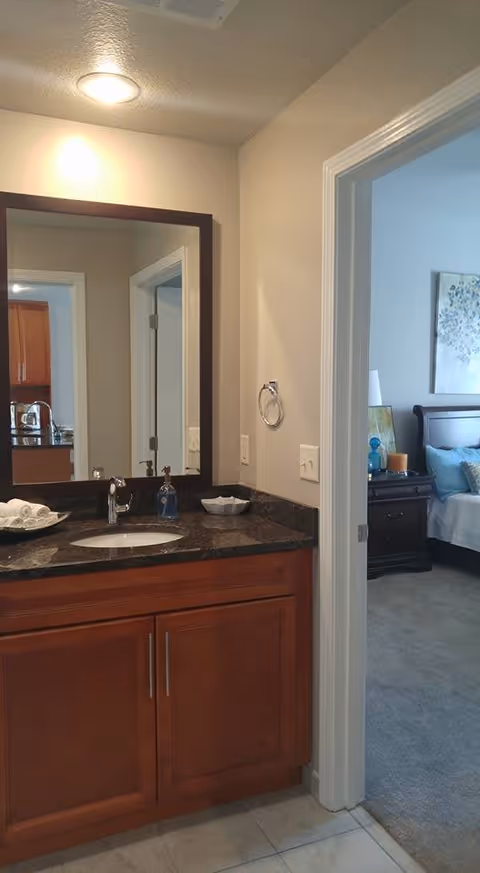 Bathroom vanity with a dark granite countertop, wooden cabinets, a sink, a soap dispenser, and a mirror reflecting a doorway and part of a kitchen. To the right, an open door leads to a bedroom with a bed, nightstand, lamp, and wall art.