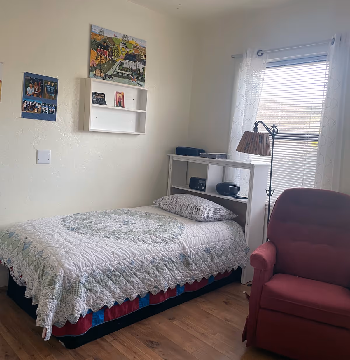 A small bedroom with a single bed covered by a quilt, a white shelving headboard, a floor lamp by a window, and a red armchair.