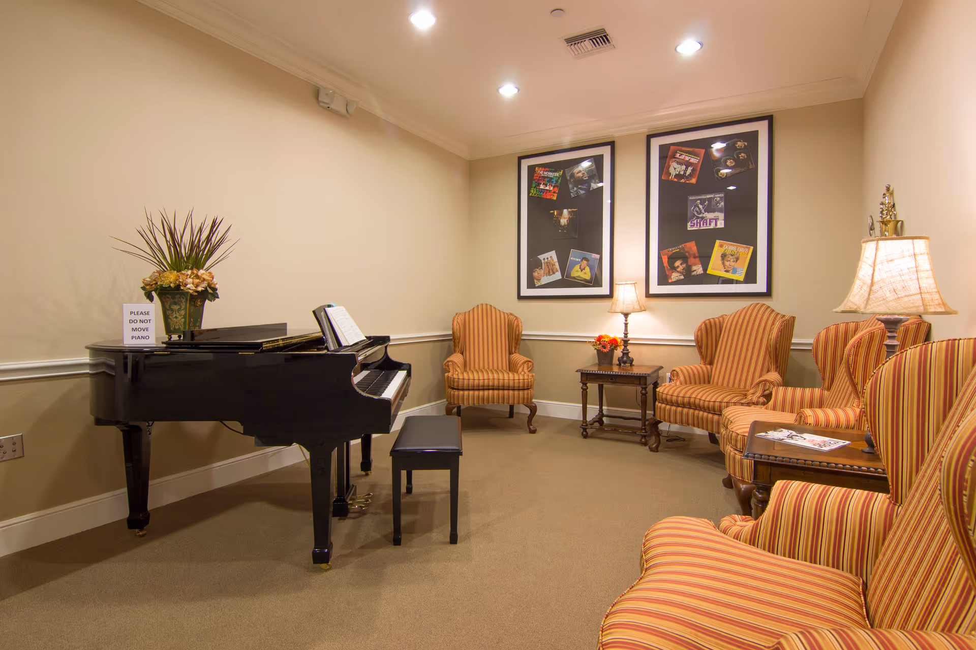A cozy sitting room with a black grand piano on the left, a piano bench, and a sign on the piano that reads 'PLEASE DO NOT MOVE PIANO'. The room has beige walls and carpet, with four striped armchairs arranged around two small wooden side tables. Two framed pictures with album covers hang on the wall above a table lamp and a small flower arrangement.