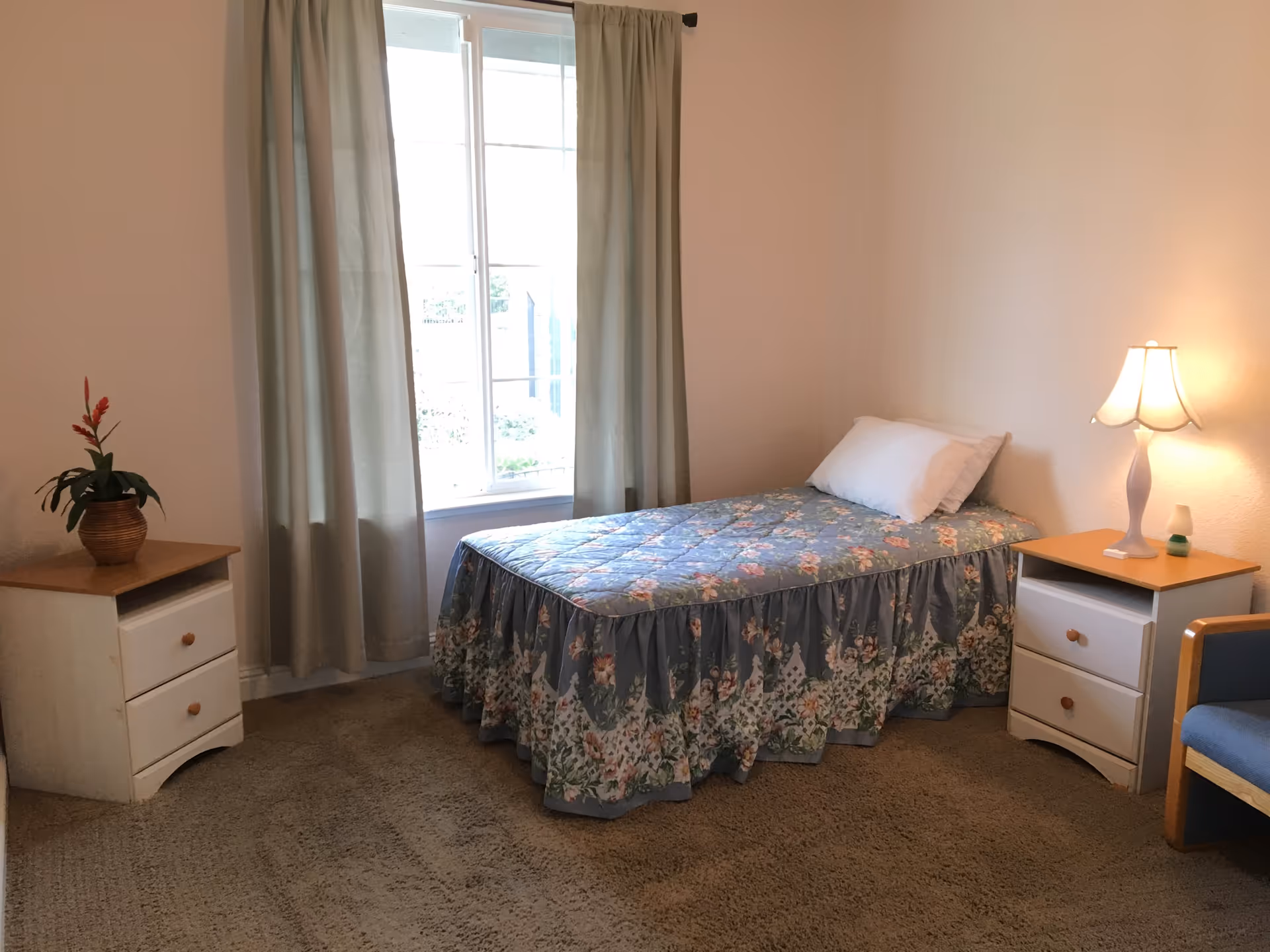 A simple bedroom with a single bed covered in a floral bedspread, a white pillow, a wooden nightstand with a lamp, another nightstand with a potted plant, a window with light-colored curtains, and a blue armchair.