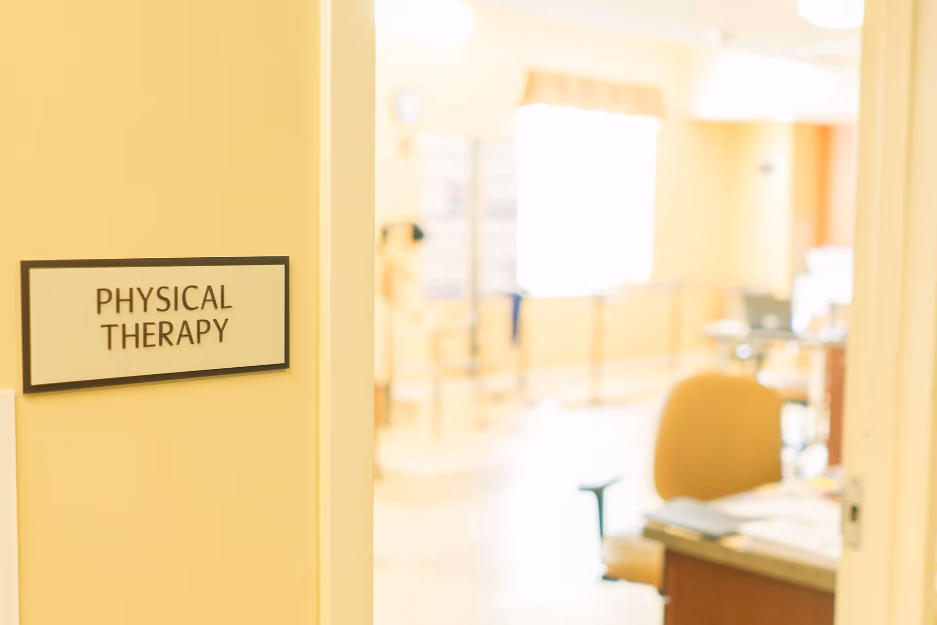 View of a physical therapy room entrance with a sign on the wall reading 'PHYSICAL THERAPY'. Inside the room, there is a desk with papers and an office chair, with bright natural light coming through a window in the background.