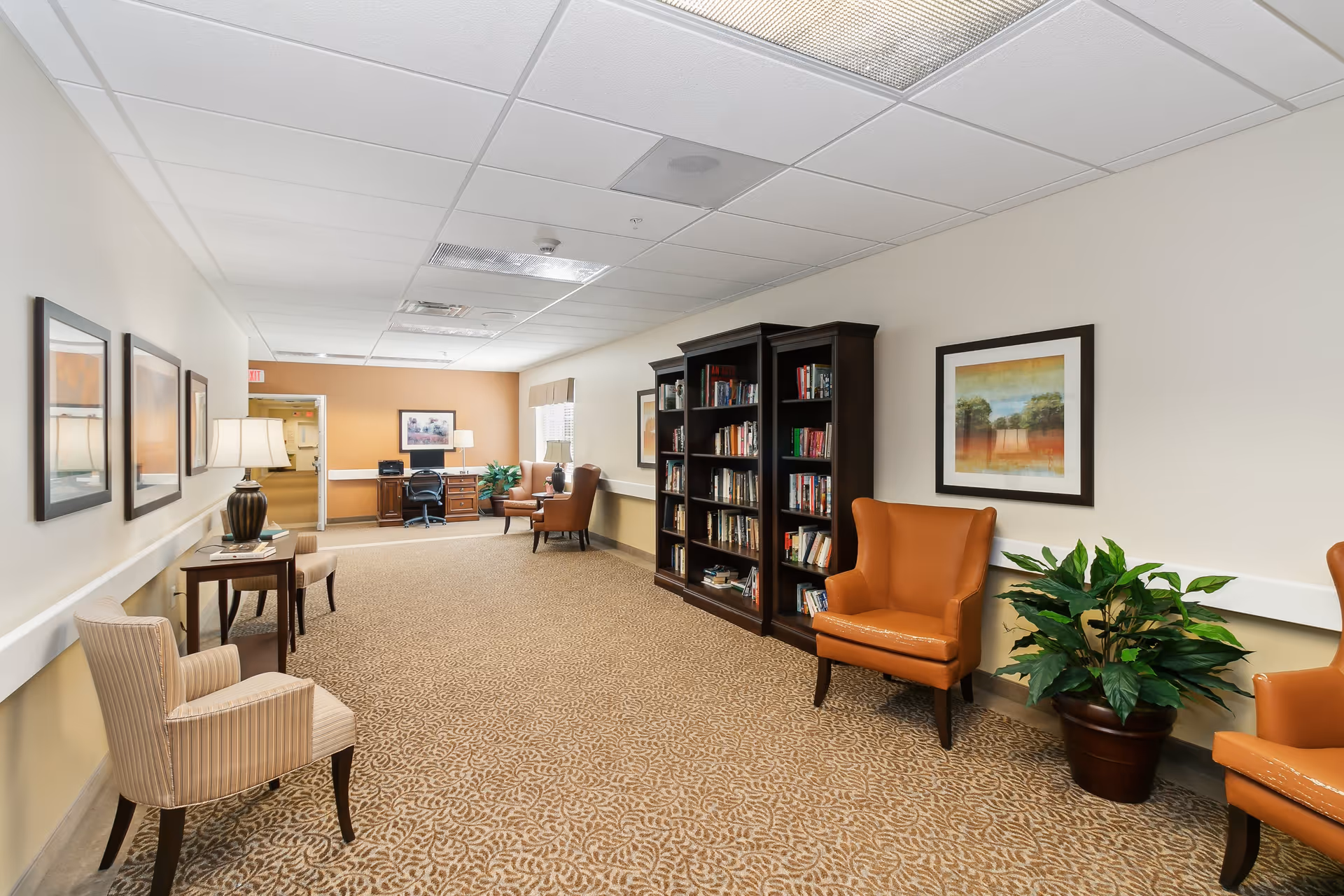 A well-lit interior hallway area in a senior living facility featuring patterned carpet, beige walls, and a white ceiling with fluorescent lights. The space includes several chairs, a small table with a lamp, framed artwork on the walls, a large bookshelf filled with books, and a potted plant. At the far end, there is a desk with a chair and additional seating near a window.