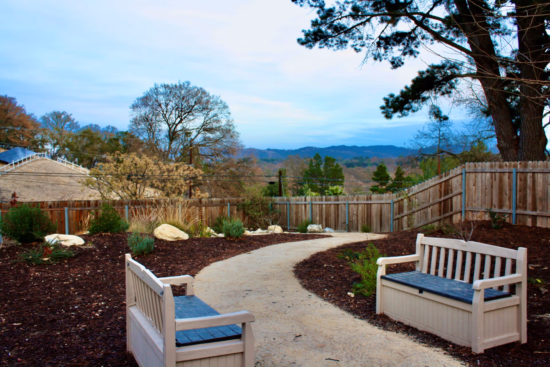 Two wooden benches beside a winding gravel path in a fenced garden with trees and distant hills.