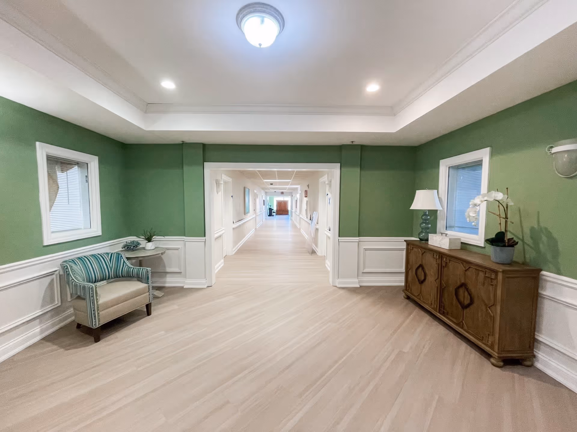 A wide hallway in a senior living facility with green walls and white wainscoting. There is a striped armchair and small round table with a plant on the left side, and a wooden cabinet with a lamp and orchid on the right side. The hallway extends into the distance with doors and handrails along the walls.